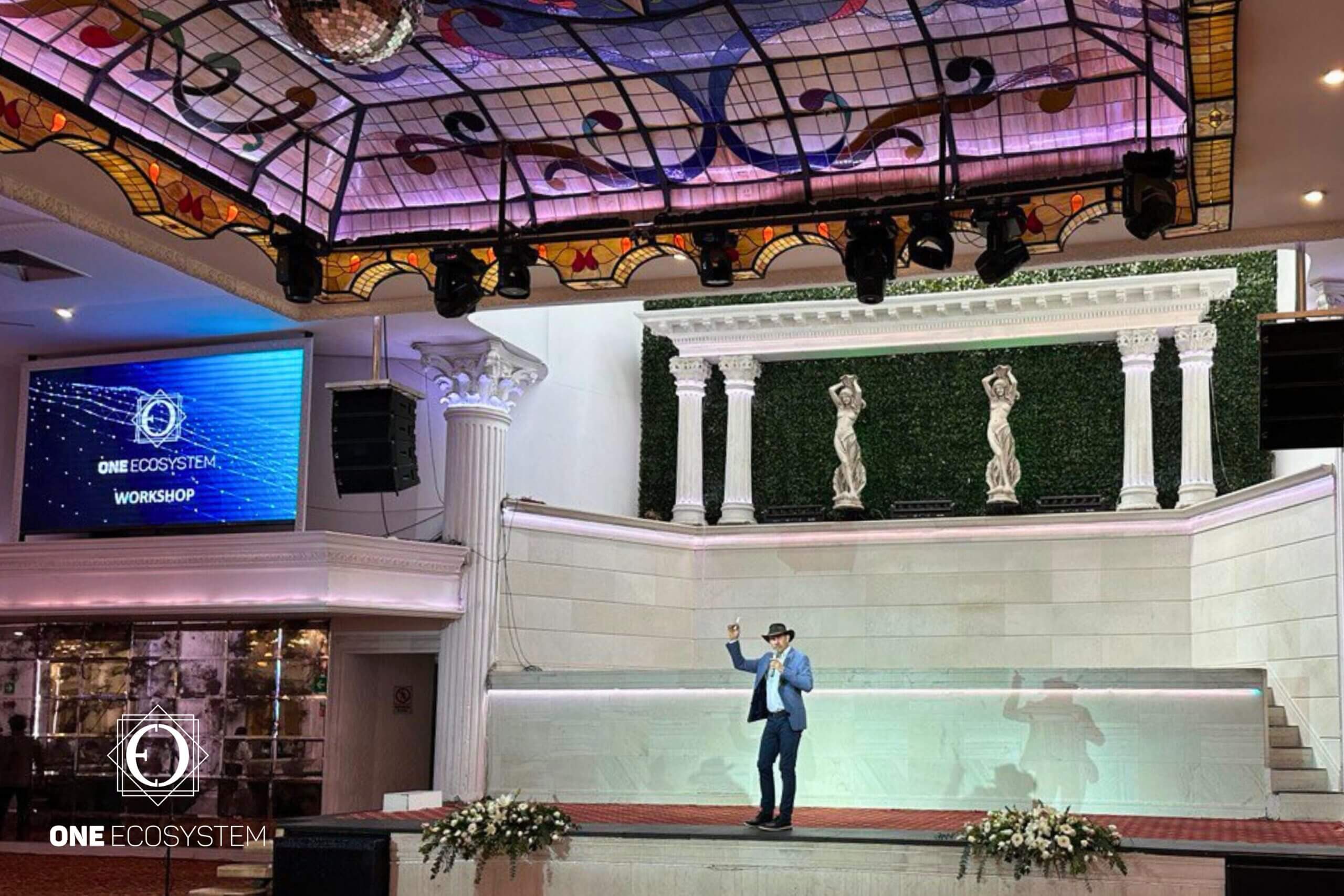 Isang taong nakatayo sa entablado na may hawak na mikropono at nagsenyas gamit ang kanyang mga kamay. Sa likod nito ay may pinalamutian na background at kisame. SA KALIWANG SCREEN, MAKIKITA MO ANG "ONE ECOSYSTEM WORKSHOP" NA MAY MGA HIGHLIGHT TUNGKOL SA MGA CRYPTOCURRENCY AT PAGSASANAY SA NETWORK.