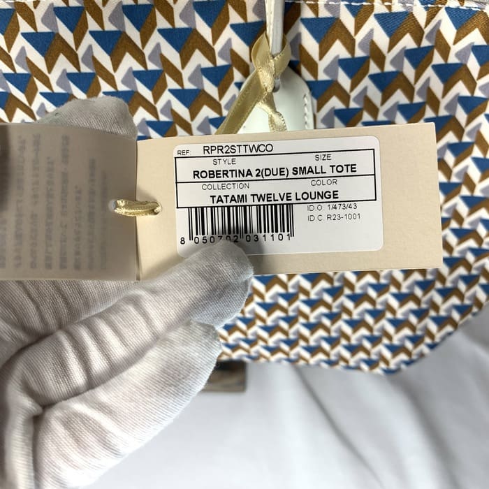 Ang guwantes na kamay ay humahawak ng tag ng produkto na "ROBERTINA 2 (DUE) SMALL TOTE" sa tuktok ng tela ng pattern ng tatami na may isang geometric pattern sa asul, kayumanggi at puti, na kung saan ay ang estilo ng lagda ng maliit na koleksyon ng tote ni ROBERTA PIERI.