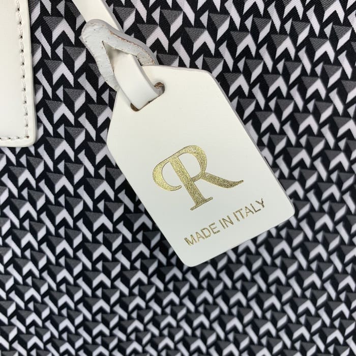 Ang isang puting katad na ROBERTA PIERI tag na may mga titik na "R" sa ginto at ang mga salitang "MADE IN ITALY" ay nakabitin mula sa background ng isang itim at puting geometric na tela.