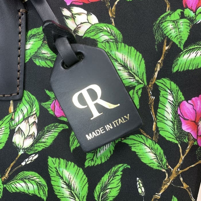 Ang itim na ROBERTA PIERI luggage tag na may gintong "R" at tekstong Italyano ay nasuspinde mula sa isang floral na tela na may berdeng dahon at kulay-rosas na bulaklak, na umakma sa mini duffel na may walang-hanggang kagandahan ng Italyano.