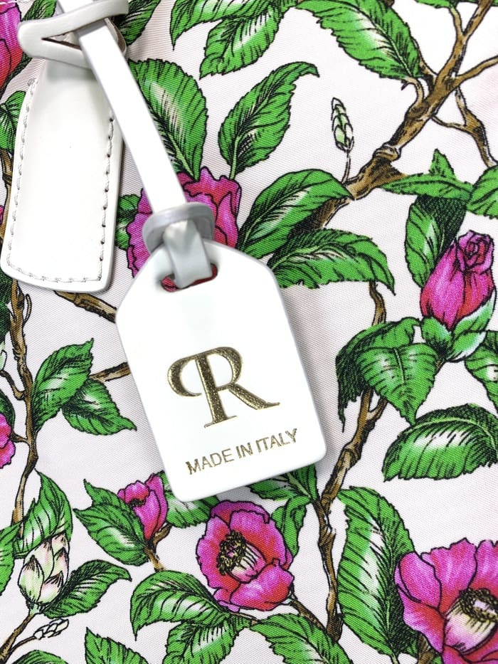 Close-up ng ROBERTA PIERI mini duffel bag. peduncles na may berdeng dahon at kulay-rosas na bulaklak; Ang puting tag na naka-attach sa 2-way bag ay nagtatampok ng isang gintong logo na "R" at ang mga salitang "MADE IN ITALY."